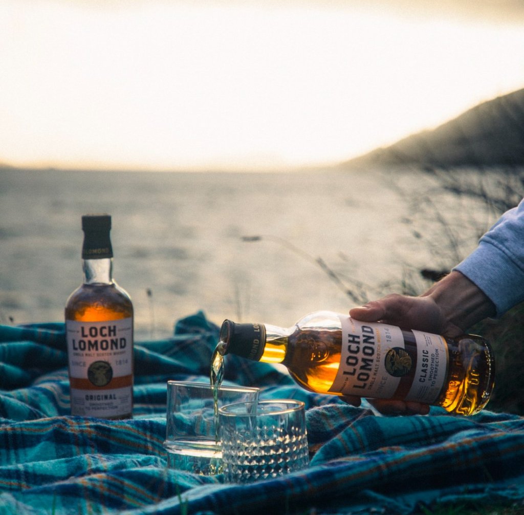 Man pouring Loch Lomond whiskey into glasses on a picnic blanket in front of Loch Lomond.