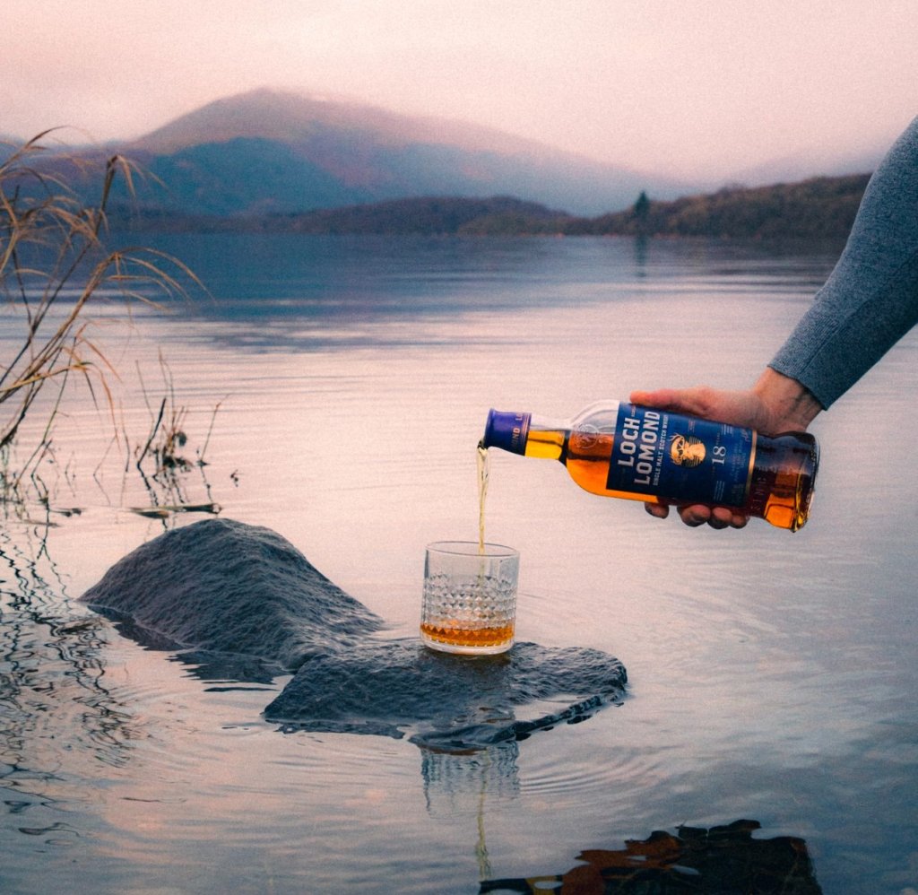 Man pouring whiskey into a glass on a stone in Loch Lomond.