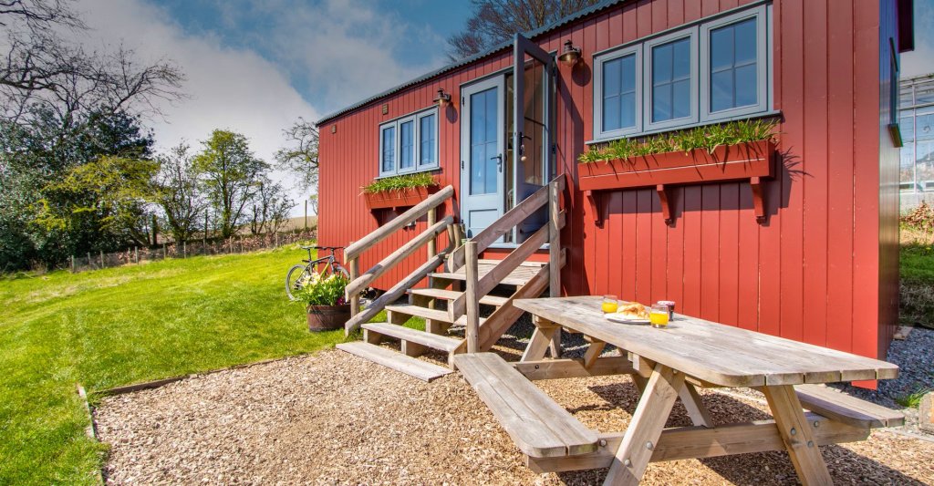 Two glasses of orange juice and pastries are sitting on an outdoor picnic table in front of the Bankhead Shepherds Hut.