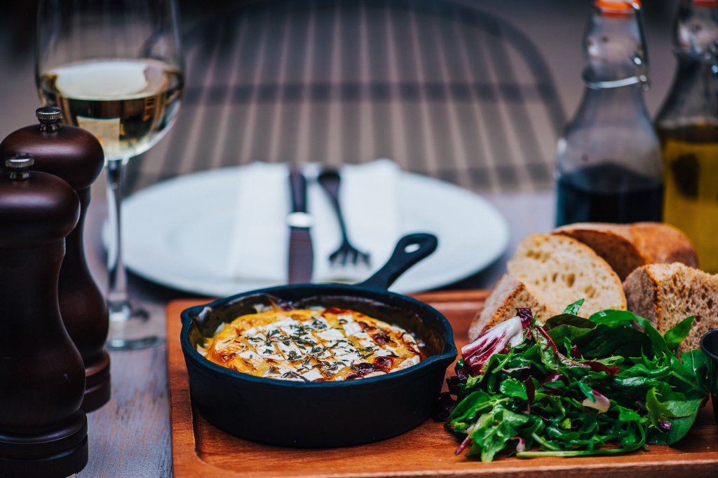 a skillet containing a baked camembert on a wooden tray with a rustic bread and fresh salad. It appears to be a very premium restaurant.