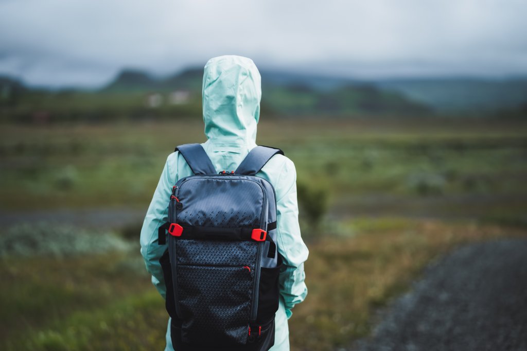 A woman in a raincoat with her hood up, looking at the landscape in the background. She is wearing a backpack.